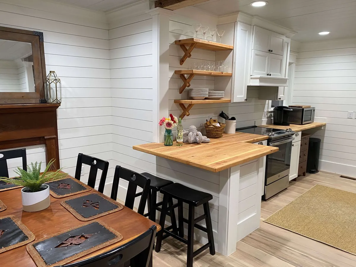 The image presents a warm dining area adjacent to a kitchen island, with open shelving and a fireplace adding charm and comfort. The setup is ideal for shared meals, entertaining, and morning routines, embodying the space's welcoming spirit.