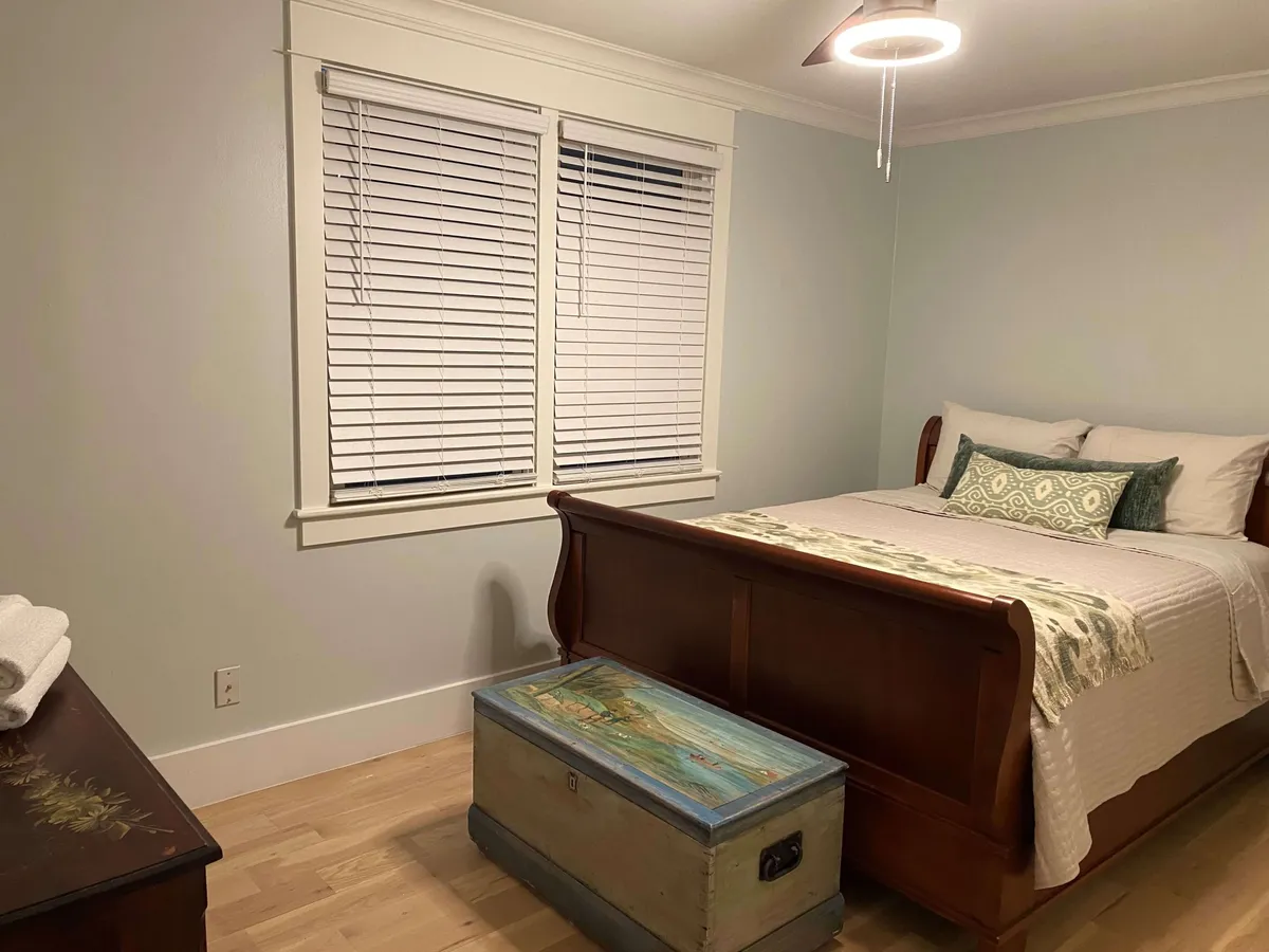 A serene bedroom with a plush bed topped with decorative pillows and warm blankets. The foot of the bed features a painted trunk, and a window with blinds allows for both natural light and privacy. The space exudes coziness and calm, perfect for relaxation and restful sleep.