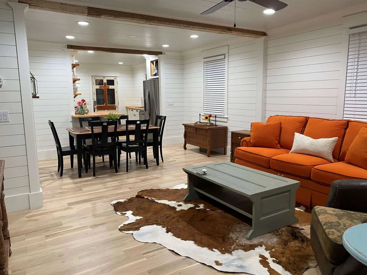 This image captures a vibrant, open-concept living and dining space featuring a standout orange sofa, a rustic wooden dining table, a cowhide rug, and a green coffee table. The room is flooded with natural light, highlighting the modern furnishings and inviting layout perfect for gatherings and relaxed activities.