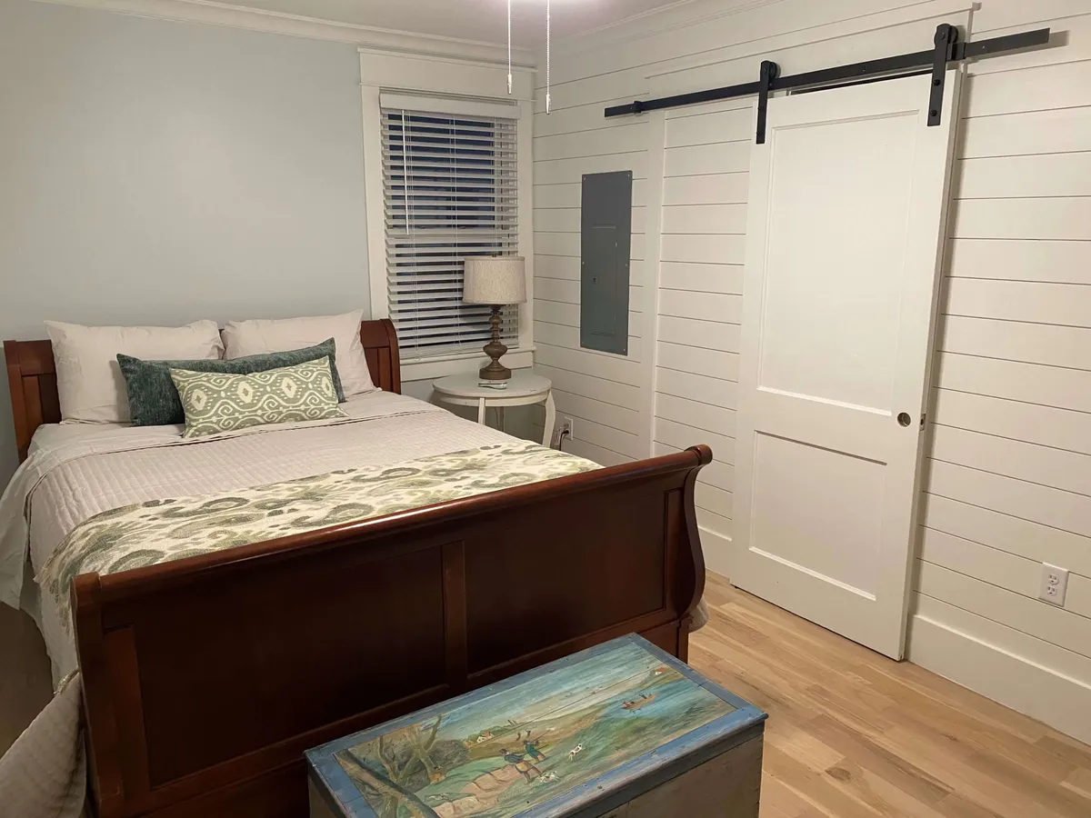 A stylish bedroom with a wooden bed frame, inviting throw pillows, and a lamp on the nightstand. A sliding barn door adds a modern touch, while a decorative chest at the bed's end offers storage. The overall mood is relaxing, perfect for unwinding or quiet moments like reading or sipping tea.